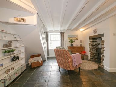 A sitting room with a striped armchair a stone fireplace and shelves by a window at Blacksmiths Cottage in Crantock
