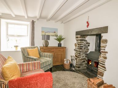 A living room with a wood burning stove stone fireplace armchairs a wooden cabinet and a window at Blacksmiths Cottage in Crantock