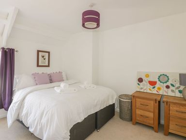 A bedroom with a bed with white bedding and purple pillows beside wooden nightstands and a basket at Blacksmiths Cottage in Crantock