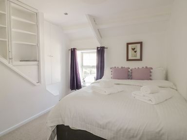 A bedroom with a bed with white linens and purple pillows next to a window with purple curtains at Blacksmiths Cottage in Crantock