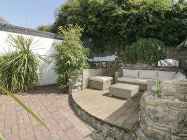 An outdoor seating area with a wicker sofa and table on a wooden deck surrounded by stone walls and greenery at Blacksmiths Cottage in Crantock