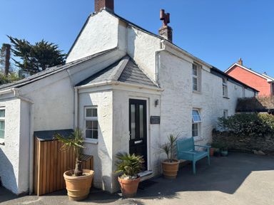 Exterior view of a white stone cottage with a black door blue bench and potted plants at Blacksmiths Cottage in Crantock