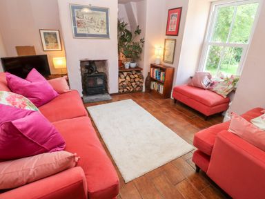 A living room with red sofas pink pillows a white rug a wood stove firewood stacked a bookshelf and a window showing greenery at Pentira in Kuggar
