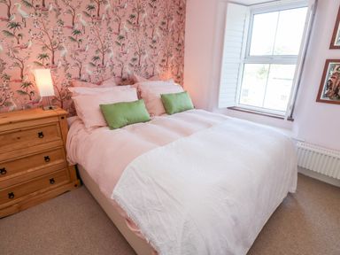 A bedroom with a double bed with pink bedding and green pillows a wooden bedside table with a lamp beside a window with white shutters at Pentira in Kuggar