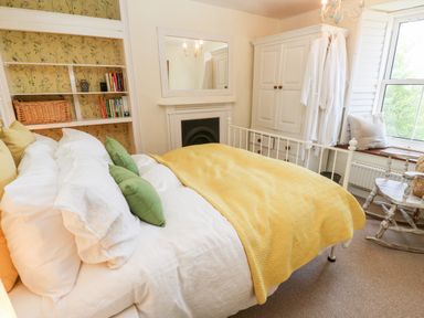 A bedroom with a bed covered in white sheets and a yellow blanket with green and yellow pillows a wooden rocking chair by a window with shutters a white wardrobe a fireplace and shelves with books and a wicker basket at Pentira in Kuggar