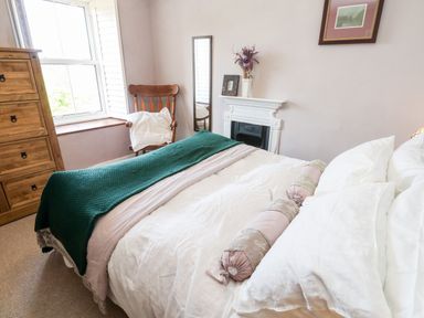 A bedroom with a bed covered in white bedding and a green blanket a wooden chair with a cushion a wooden chest of drawers a small white fireplace and a framed picture on the wall at Pentira in Kuggar