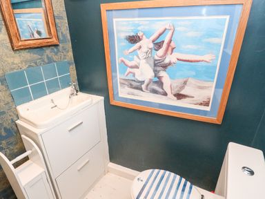 A small bathroom with a white sink cabinet blue tiled backsplash a toilet with striped seat and a framed painting on the wall at Pentira in Kuggar