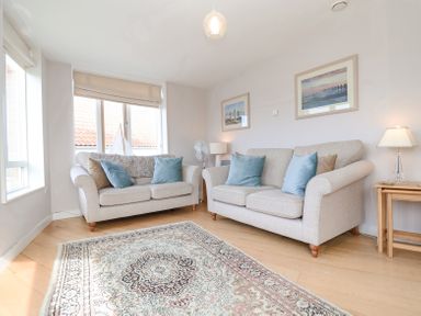 A living room with two beige sofas blue and brown cushions a patterned rug and framed pictures on the wall at 23 Tibby's Way in Southwold