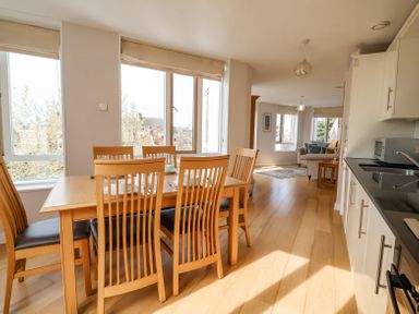 A dining area with a wooden table and six chairs next to large windows and an open living space with sofas at 23 Tibby's Way Southwold