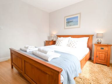 A bedroom with a wooden bed frame a blue blanket white pillows two bedside tables with lamps and a framed picture on the wall at 23 Tibbys Way in Southwold