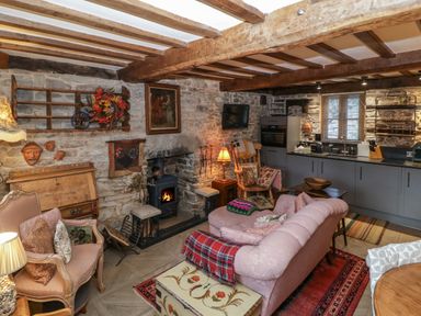 A living area with a pink sofa a wood burning stove wooden beams stone walls and a kitchen area at Hoarders Cottage in Hay-On-Wye