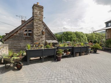 A paved outdoor area with raised planters filled with plants next to a wooden building with a stone chimney at Hoarders Cottage in Hay-On-Wye