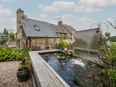 An outdoor garden with a pond with a waterfall feature next to a stone and wooden house at Hoarders Cottage in Hay-On-Wye