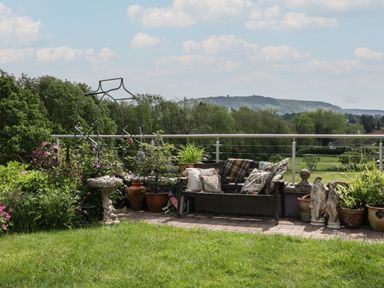 A garden seating area with a wooden bench, cushions, potted plants, and statues with a view of trees and hills at Hoarders Cottage in Hay-On-Wye