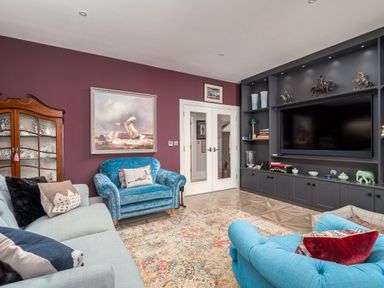 A living room with blue and gray sofas a patterned rug a painting on a purple wall and a built-in black shelving unit with a television at The Stables in Wheatley