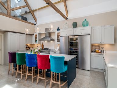 A kitchen with white cabinets and a blue island with multicolored bar stools and wooden beams at The Stables in Wheatley