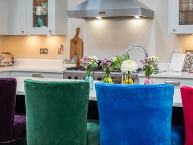 A kitchen with colorful chairs at a kitchen island and flower vases on the counter at The Stables in Wheatley