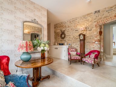 A sitting area with patterned chairs a wooden table with a lamp and flowers a large mirror and a grandfather clock at The Stables in Wheatley