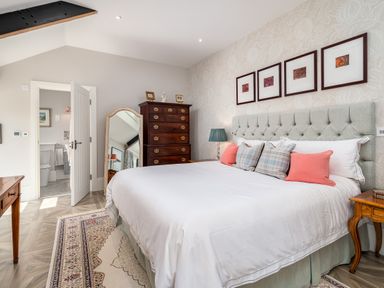 A bedroom with a large bed pillows a wooden chest of drawers a floor mirror and an open door to a bathroom at The Stables in Wheatley