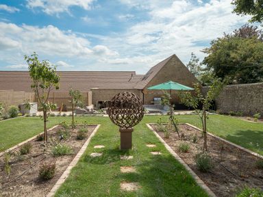 A garden with small trees flower beds and a metal spherical sculpture in front of a stone building at The Stables in Wheatley