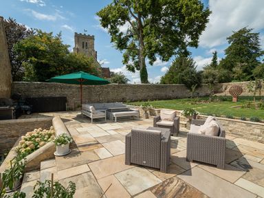 An outdoor patio with wicker chairs and a gray sofa set under a green umbrella with a stone wall and garden at The Stables in Wheatley