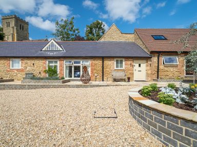 Stone cottage with gravel driveway and raised flower beds under a blue sky at The Stables in Wheatley