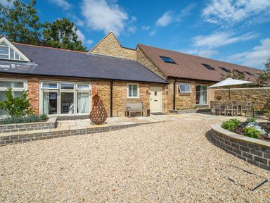 A stone cottage with gravel driveway garden sculpture bench and outdoor dining set with umbrella at The Stables in Wheatley