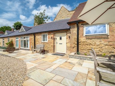 Stone exterior of a house with windows a door benches a large patio umbrella and paved patio at The Stables in Wheatley