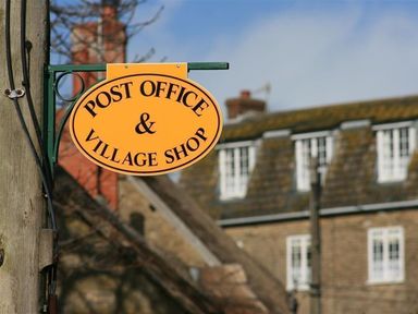 A post office and village shop sign mounted on a pole with buildings in the background
