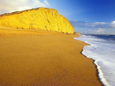 A sandy beach with waves washing ashore and a tall yellow cliff under a blue sky at Dormouse Cottage in Burton Bradstock
