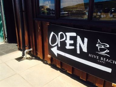A black open sign with white lettering and an arrow on a wooden wall outside a cafe at Hive Beach