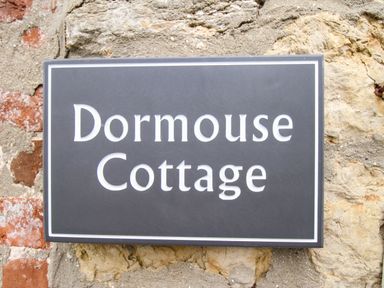 A black sign with white letters reading Dormouse Cottage on a stone wall at Dormouse Cottage Burton Bradstock