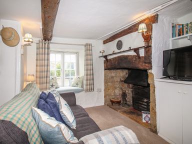 A living room with a sofa cushions a stone fireplace a bay window with curtains and a television at Dormouse Cottage in Burton Bradstock