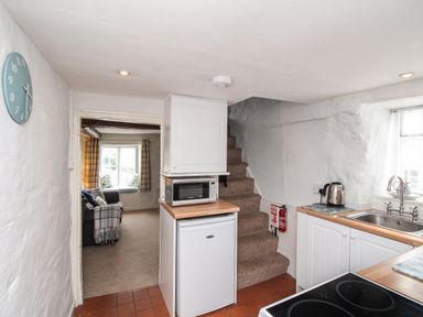 A kitchen with a microwave on a counter above a fridge with stairs beside and a living room visible through a doorway at Dormouse Cottage in Burton Bradstock