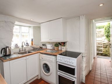 A small kitchen with white cabinets a washing machine an electric stove a stainless steel sink and a door leading outside at Dormouse Cottage in Burton Bradstock