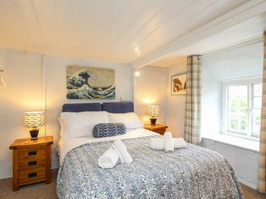 A bedroom with a bed between two wooden nightstands with lamps and a window with plaid curtains at Dormouse Cottage in Burton Bradstock
