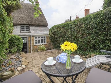 A small outdoor seating area with a round metal table holding a teapot cups and flowers in front of a thatched roof cottage surrounded by greenery at Dormouse Cottage in Burton Bradstock