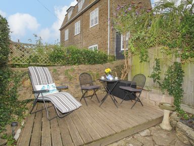 An outdoor wooden deck with a reclining chair with a cushion two folding chairs and a small round table set for tea at Dormouse Cottage in Burton Bradstock