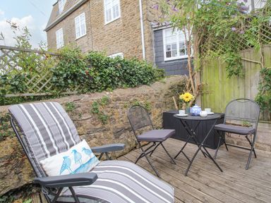 An outdoor patio area with a striped lounge chair, two black metal chairs, a small round table with a teapot, cups, and flowers at Dormouse Cottage in Burton Bradstock