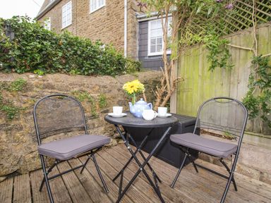 A patio area with two metal chairs and a round table with a teapot cups and flowers at Dormouse Cottage in Burton Bradstock