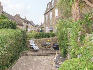 A small garden patio with chairs a table and greenery around stone houses in the background at Dormouse Cottage in Burton Bradstock