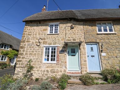 A stone cottage with two pastel-colored doors and white framed windows with flowers in front at Dormouse Cottage in Burton Bradstock