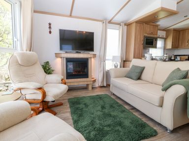A living room with two beige armchairs a beige sofa green cushions a green rug a wall-mounted TV and a fireplace at Gwel Y Mor in Nasareth near Caernarfon