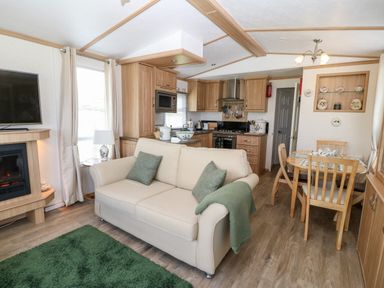 A living area with a cream sofa, green cushions, green rug, kitchen with wooden cabinets, dining table with chairs, and a fireplace at Gwel Y Mor in Nasareth near Caernarfon