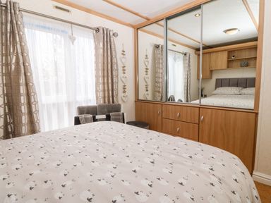A bedroom with a bed covered with patterned bedding a window with curtains a chair with towels and a wooden wardrobe with mirrored sliding doors at Gwel Y Mor Nasareth near Caernarfon