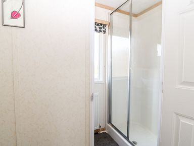 A bathroom with a glass-enclosed shower, window, radiator, framed artwork on the wall, and a gray bath mat at Gwel Y Mor in Nasareth near Caernarfon