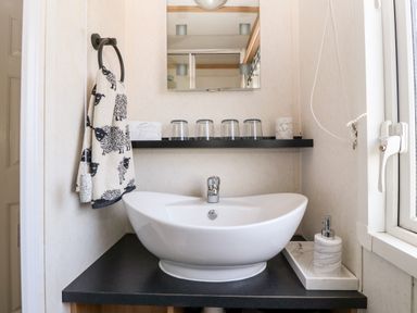 A bathroom sink with a towel hanging on a ring and cups on a shelf above the sink at Gwel Y Mor in Nasareth near Caernarfon