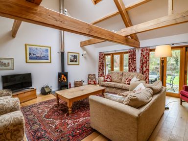 A living room with wooden beams a wood burning stove sofas a wooden coffee table and glass doors opening to a garden at Bridge Cottage in Dalwood