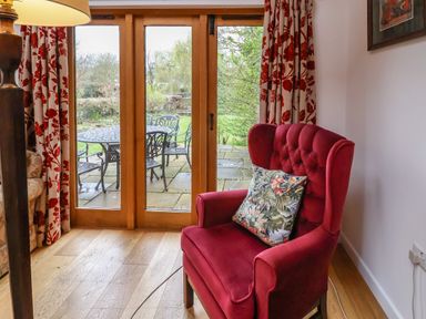 A red armchair with a patterned cushion by wooden framed glass doors showing a patio with metal table and chairs at Bridge Cottage in Dalwood