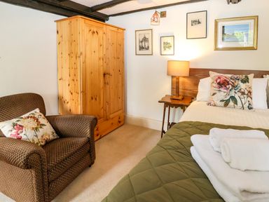 A bedroom with a bed with folded towels on the blanket a wooden wardrobe armchair with a floral pillow and a bedside table with a lamp at Bridge Cottage in Dalwood